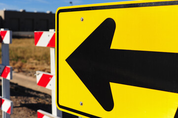 Close-up of yellow directional traffic sign with left-pointing arrow, positioned in front of...