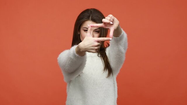 Serious brunette woman in fluffy white sweater focusing at camera through photo frame gesture, cropping view, capturing moment, concentrating at target. Indoor isolated on orange background