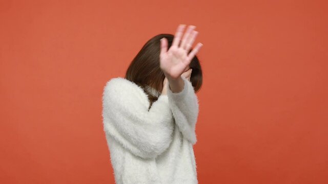 I don`t want to look at this. Scared confused brunette woman in fluffy white sweater covering eyes with hand in shock, hiding, asking to stop. Indoor studio shot isolated on orange background