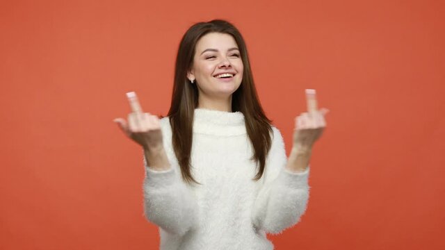 Carefree rude woman in white fluffy sweater showing middle fingers and smiling, having fun and relaxing to all problems and negativity. Indoor studio shot isolated on orange background