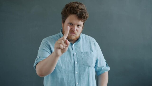 Portrait of strict young man waving finger demanding decent behavior looking at camera with serious face standing alone against gray background