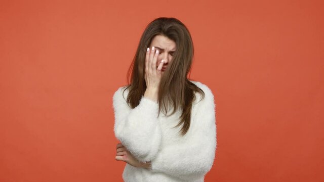 Upset worried brunette woman in fluffy white sweater closing face with hand making facepalm gesture, ashamed and depressed, making huge mistake. Indoor studio shot isolated on orange background