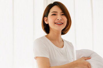 Adult beautiful asian woman pretty with short brown hair and wearing white t-shirt holding white pillow smiling and happy casual in morning on one day