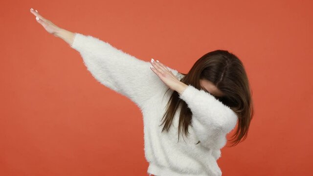 Cheerful positive brunette woman in fluffy white sweater dancing and making dub gesture, dabbing raising arms and covering eyes, having fun. Indoor studio shot isolated on orange background