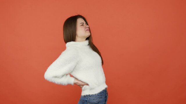 Exhausted brunette woman in white fluffy sweater touching injured sore back, suffering spasms from torn muscles, pinched nerve, kidney stones. Indoor studio shot isolated on orange background