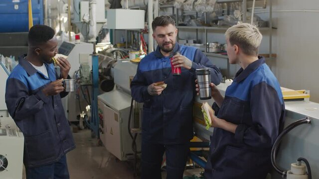  Medium shot of male and female plant workers in uniforms chatting and eating sandwiches beside manufacturing equipment in workplace