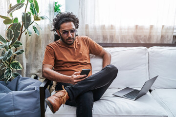 Portrait of a dressed in casual clothing indian guy he sits at home on sofa and spends his holiday using telephone and laptop.