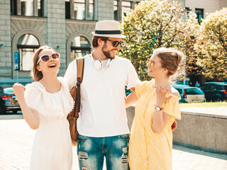 Obraz premium Group of young three stylish friends posing in the street. Fashion man and two cute female dressed in casual summer clothes. Smiling models having fun in sunglasses.Cheerful women and guy outdoors