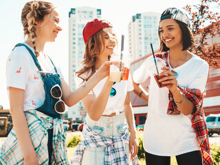 Three young beautiful smiling hipster female in trendy summer clothes.Sexy carefree women posing outdoors.Positive models holding and drinking fresh cocktail smoothie drink in plastic cup with straw