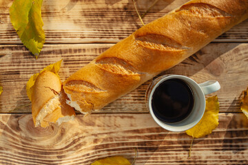 A fresh, crisp baguette, broken in two, lies on a wooden background among the foliage. Next to it is a mug of brewed coffee
