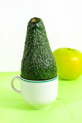 A whole green avocado stands in a mug with an apple in the background.