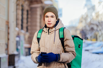 Handsome delivery man wearing warm clothes in winter city street. Teenager courier with green backpack outdoors.