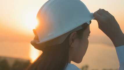 a woman engineer is putting a protective helmet on her head at sunset.