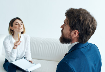 A man in a blue jacket and a beard talks to a woman on the sofa indoors