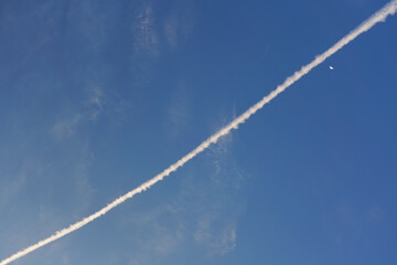 Airplane white pink condensation trail (also known as chemtrails in conspiracy theory) painted diagonally over clear morning sky, moon near