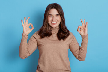 Portrait of excited young attractive girl looking directly at camera with charming smile, showing ok signs with both hands, wearing beige turtleneck, posing isolated over blue background.