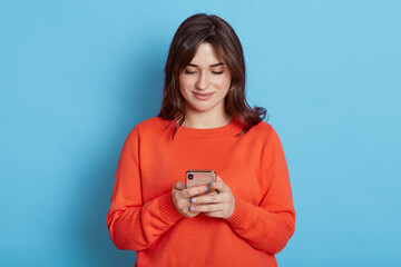 Pretty European woman with dark hair, watches video on smart phone isolated on blue wall, sends...