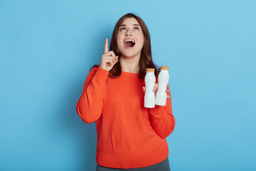 Young dark haired woman holding two bottles of milk, has idea or question pointing finger with happy and exited face, looking up, lady wearing casual shirt, keeping healthy diet.