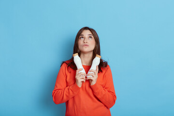 European young and healthy Caucasian woman holding two bottles fresh milk, looking up with pensive facial expression, lady with yogurt wearing casual orange sweater isolated over blue background.