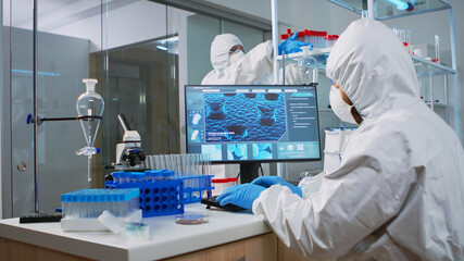 Naklejka premium Chemist man in coverall typing on computer analysing vaccine evolution in modern equipped laboratory. Team of doctors in ppe suit using high tech for researching treatment against covid19 virus