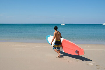 Obraz premium A sportman man with paddleboard going to the sea having fun and enjoying their vacations outdoors at the beach of Thailand. Surfer lifestyle. SUP or Stand Up Paddle Boarding.