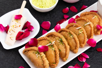 Traditional Indian Holi Celebration - Gujia Or Gujiya Mithai Made In Desi Ghee Pista Badam Served In Tray With Cold Kulfi. Gulal And Rose Petal Floral Decoration On Black Background