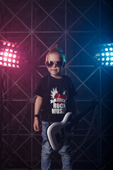 Little boy in sunglasses with a guitar on a background of colored lights. The inscription on the T-shirt Rock music. Hobby and leisure concept.