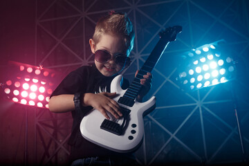Little boy in sunglasses with a guitar on a background of colored lights. The inscription on the T-shirt Rock music. Hobby and leisure concept.