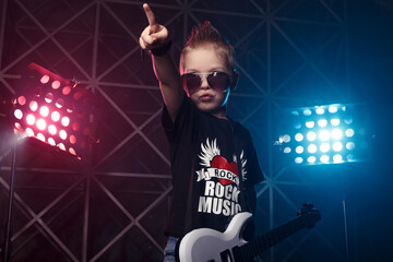 Little boy in sunglasses with a guitar on a background of colored lights. The inscription on the T-shirt Rock music. Hobby and leisure concept.