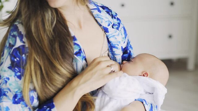 Faceless young mom in blue pajamas breastfeeds baby and fixes long loose flowing hair Close-up woman sitting on bed in bedroom at home