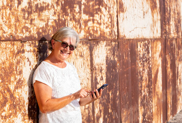 Beautiful senior woman standing outdoor close to a rusty metal panel using her mobile phone, smiling. Sunny summer day