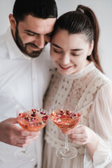 Smiling romantic caucasian couple celebrating holiday with glasses of cold sparkling champagne cocktail with rose petals and drinking straws together. Wedding anniversary, Valentines Day