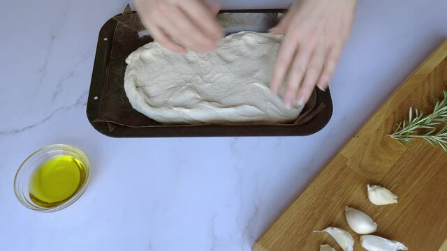 Top down view of person at home baking focaccia bread on white marble surface with raw ingredients in the frame. Person kneads and presses dough into shape.