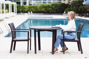 Young female freelancer sitting near the pool with her laptop in the hotel. Distant work concept