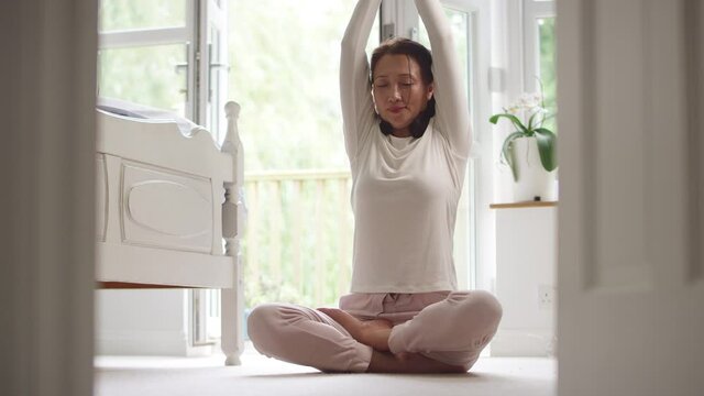 Mature Asian woman in pyjamas sitting on bedroom floor meditating in yoga pose - shot in slow motion