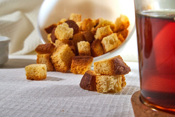 Square toasted pieces of homemade delicious rusk, hardtack, Dryasdust, zwieback on a plate and black tee in a cap on a white tablecloth.