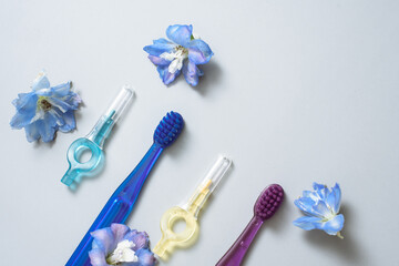 Items for cleaning teeth on a light gray background. Colored toothbrushes and a brush for cleaning between teeth next to fresh flowers