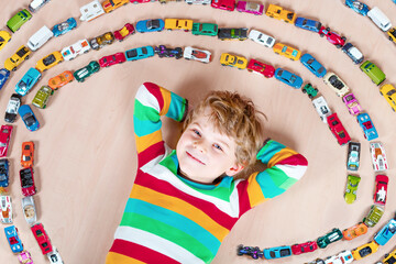 Cute little blond kid boy playing with lots of toy cars indoor. Child wearing colorful shirt. Happy preschooler having fun at home or nursery. Big collection of different vehicles. Happiness game.