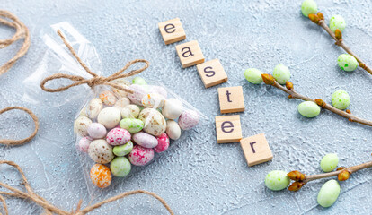 Fototapeta na wymiar Chocolate Easter eggs in glaze in a transparent bag, branches of a spring tree and wooden letters on a light blue background. Happy Easter time. Preparation for holiday, gift wrapping. Banner.