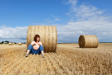 Mature woman relaxes in a harvested corn field on a sunny autumn day. She is sitting against a hay bale and is wearing casual clothing.  - Powered by Adobe