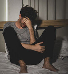 stressed and upset young caucasian man dressed casually sitting on a bed hand holding head....