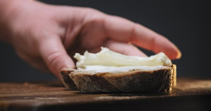 Woman spreads fresh soft cheese on rye bread slice with shiny knife on wooden cutting board in kitchen extreme close view