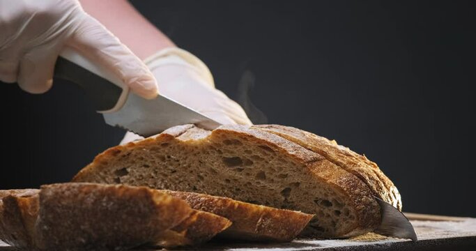 Person in white gloves cuts slices of fresh hot rye bread loaf with sharp knife on wooden board on black background closeup