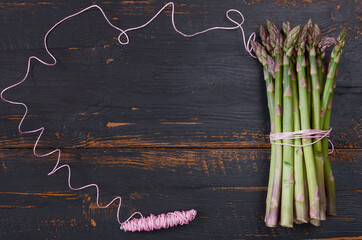 Spring frame with pink thread and green asparagus bunch on the black wooden background. Healthy vegetables for fresh salad ready for cooking . Top view