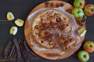 Traditional sliced apple pie with spices on the brown wooden board. Apple pie decorated with fresh apples, carob pods, anise stars and cinnamon sticks  on the black background. Top view 