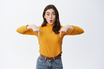 Candid woman looking surprised and interested, asking to check out advertisement, pointing fingers down and say wow amazed, standing on white background