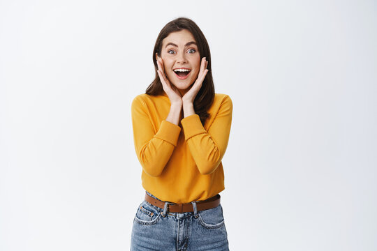 Wow amazing. Impressed young woman smiling excited, holding hands on face with natural light make up, staring at camera astonished, see awesome advertisement, white background