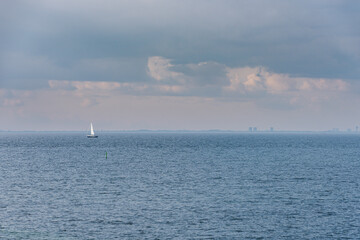 Fototapeta premium A lone sailing boat at sea off Malmo in the Oresund Strait with Copenhagen skyline and a cloudy sky suggests relaxation or break in calm water. Sailboat in the ocean conveys tranquillity and serenity
