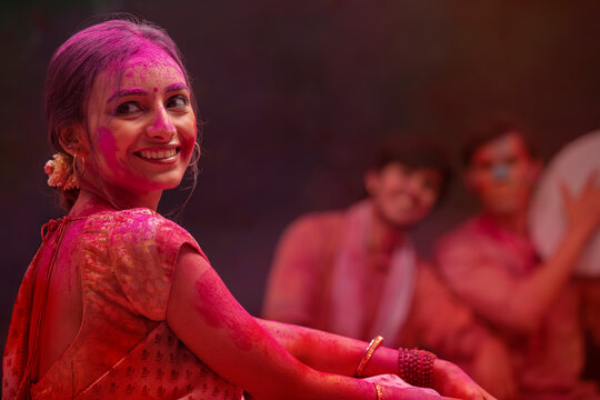 A HAPPY YOUNG WOMAN TURNING AROUND AND LOOKING AWAY DURING HOLI CELEBRATIONS WITH FRIENDS ENJOYING BEHIND	