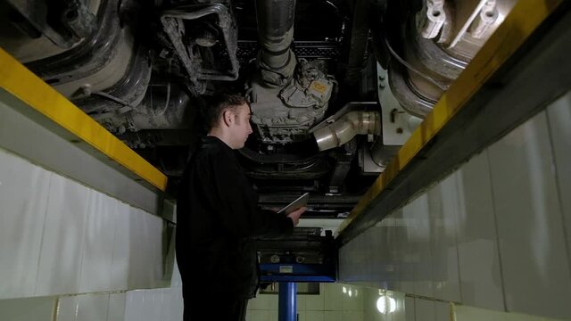 Car mechanic in  uniform makes an inspection of truck at service station. inspector records all breakdowns and problems with car in modern tablet. engineer works under car lifting machine in garage.